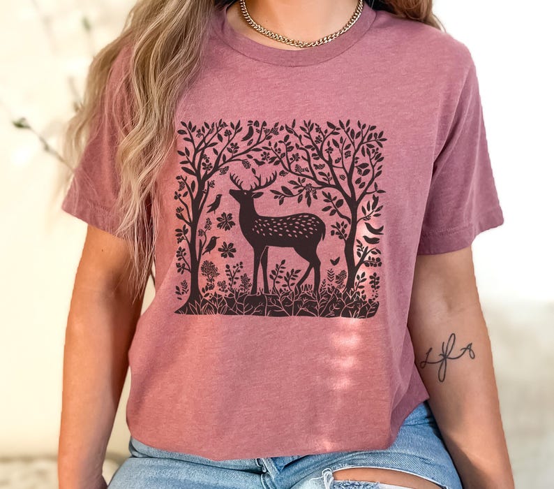 K&ouml;nnte beinhalten: Ein mauvefarbenes T-Shirt mit einem schwarz-wei&szlig;en Grafikdruck eines Hirsches in einer Waldszene. Die Grafik zeigt einen Hirsch mit Geweih, B&auml;ume und V&ouml;gel.