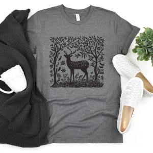 K&ouml;nnte beinhalten: Ein dunkelgraues T-Shirt mit einem schwarz-wei&szlig;en Grafikdruck eines Hirsches, der in einem Wald steht. Die Grafik zeigt B&auml;ume, Bl&auml;tter und V&ouml;gel.