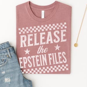 May include: A dusty rose t-shirt with the text "RELEASE the EPSTEIN FILES" in white, framed by a checkered pattern. The shirt is folded, with a pair of blue jeans and a gold necklace visible.