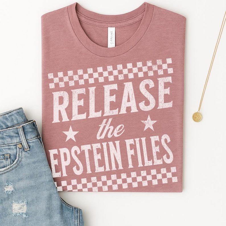 May include: A dusty rose t-shirt with the text "RELEASE the EPSTEIN FILES" in white, framed by a checkered pattern. The shirt is folded, with a pair of blue jeans and a gold necklace visible.