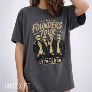 May include: Dark gray unisex t-shirt featuring a graphic of four historical figures wearing sunglasses, with the text "FOUNDERS TOUR" and the dates "1776-2026" in gold. The shirt has a vintage, worn look.