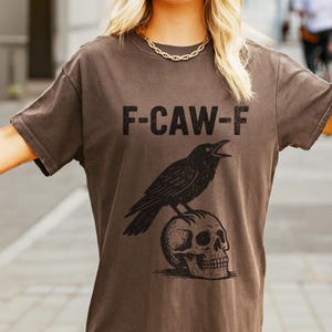 May include: A dark brown t-shirt with the text "F-CAW-F" and a black illustration of a crow perched on a skull. The crow is depicted with its beak open. The shirt has short sleeves.