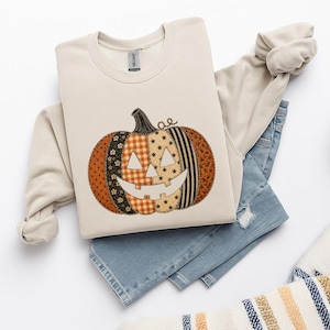 May include: A beige crewneck sweatshirt featuring a large pumpkin graphic composed of various fabric-like patterns including orange plaid, black polka dots, and star prints. The pumpkin has a carved face with triangular eyes and a wide grin.