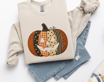 May include: A beige crewneck sweatshirt featuring a large pumpkin graphic composed of various fabric-like patterns including orange plaid, black polka dots, and star prints. The pumpkin has a carved face with triangular eyes and a wide grin.