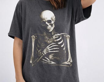 May include: A charcoal gray short-sleeve t-shirt featuring a graphic print of a human skeleton holding a small cat. The skeleton is positioned centrally on the chest, with its bony hands cradling the cat against its ribcage. The shirt has a faded, washed-out appearance.