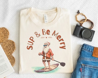Könnte beinhalten: Ein cremefarbenes T-Shirt mit einem Aufdruck von Weihnachtsmann mit Sonnenbrille und rot-weißem Anzug beim Paddelsurfen. Der Text "SUP & Be Merry" befindet sich in einem Kreis um den Weihnachtsmann.