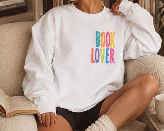 May include: A white long-sleeve crewneck sweatshirt featuring the text BOOK LOVER printed in a vertical, multi-colored sans-serif font on the upper left chest area. The letters are colored in shades of blue, orange, pink, yellow, and purple.