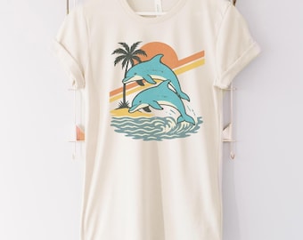 Może przedstawiać: Kremowy t-shirt z grafiką retro przedstawiającą dwa turkusowe delfiny wyskakujące z fal oceanu. Palmę i zachodzące słońce z pomarańczowymi i żółtymi paskami dopełniają projekt. Koszulka ma krótkie rękawy i okrągły dekolt.