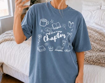 May include: A blue short-sleeve t-shirt featuring white line art illustrations of books, a coffee mug, a crown, flowers, and glasses. The text "Just One more Chapter" is printed in the center in a white script and sans-serif font combination.