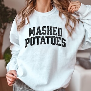 Puede incluir: Sudadera blanca con la frase "MASHED POTATOES" impresa en letras negras y gruesas. El texto está centrado en la parte delantera de la sudadera. La prenda tiene mangas largas.