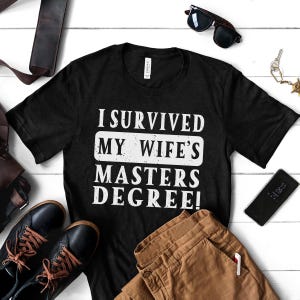 Peut inclure: T-shirt noir avec un texte blanc qui dit "J'ai survécu au master de ma femme !"