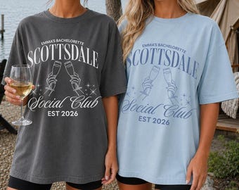 May include: Two oversized t-shirts, one charcoal gray and one light blue, each featuring white or dark blue text that reads Emma's Bachelorette Scottsdale Social Club EST 2026, accompanied by a graphic of two hands holding champagne flutes.