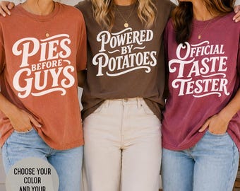 May include: Three short-sleeve crew neck t-shirts in terracotta, brown, and maroon. The terracotta shirt features white text reading Pies Before Guys. The brown shirt features white text reading Powered By Potatoes. The maroon shirt reads Official Taste Tester.