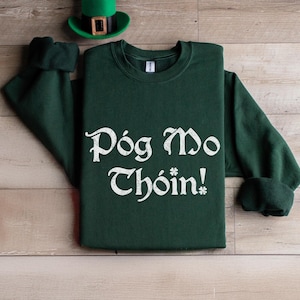 May include: A dark green sweatshirt with the text "Póg mo Thóin!" in white lettering. The text is in a vintage style font.