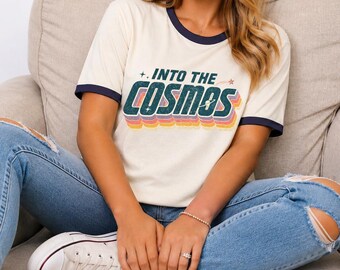 May include: A cream-colored ringer t-shirt with dark blue trim on the collar and sleeves. The front features the text "INTO THE COSMOS" in a retro, multi-layered font with green, orange, and purple colors, accented by small star and planet icons.