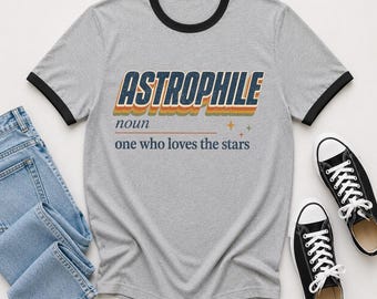 May include: A heather gray ringer t-shirt with black trim on the collar and sleeves. The chest features the text ASTROPHILE in a retro multi-colored font, followed by the definition noun, one who loves the stars, and small star icons.