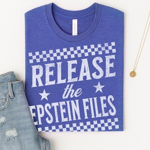 May include: A blue t-shirt with the text "RELEASE the EPSTEIN FILES" in white, with a checkered pattern above and below the text. The shirt is paired with blue jeans and a gold necklace.