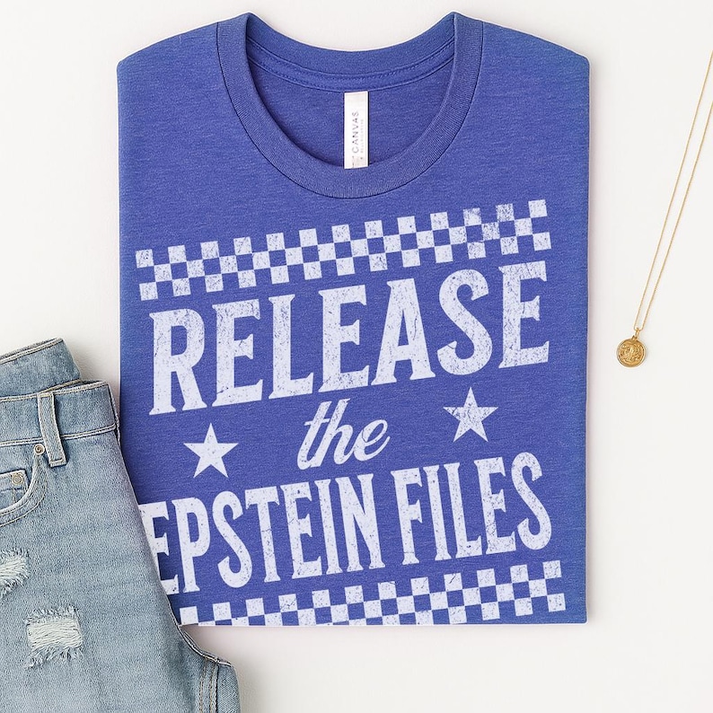 May include: A blue t-shirt with the text "RELEASE the EPSTEIN FILES" in white, with a checkered pattern above and below the text. The shirt is paired with blue jeans and a gold necklace.