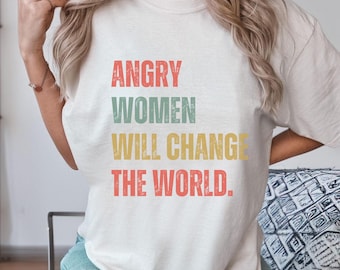 May include: Cream-coloured t-shirt with the text "ANGRY WOMEN WILL CHANGE THE WORLD" in red, green, yellow, and red lettering. The shirt has a vintage, slightly faded look.
