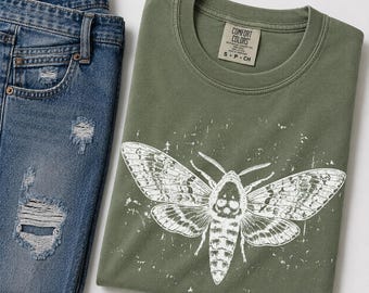 May include: An olive green crew neck t-shirt featuring a white distressed graphic of a moth with a skull pattern on its thorax. The shirt has a Comfort Colors brand label at the neck. Beside the shirt are blue denim jeans with distressed rips.
