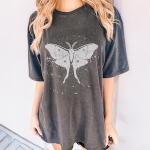May include: Dark grey t-shirt featuring a large, distressed white moth graphic. The moth design has detailed wing patterns and a vintage aesthetic. The shirt has short sleeves and a relaxed fit, suitable for casual wear.