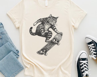 Op de afbeelding: Crèmekleurig T-shirt met een zwart-wit illustratie van een kat die skateboard. De kat is in de lucht, balancerend op het skateboard. Een paar zwarte sneakers met witte zolen en veters zijn rechts zichtbaar.