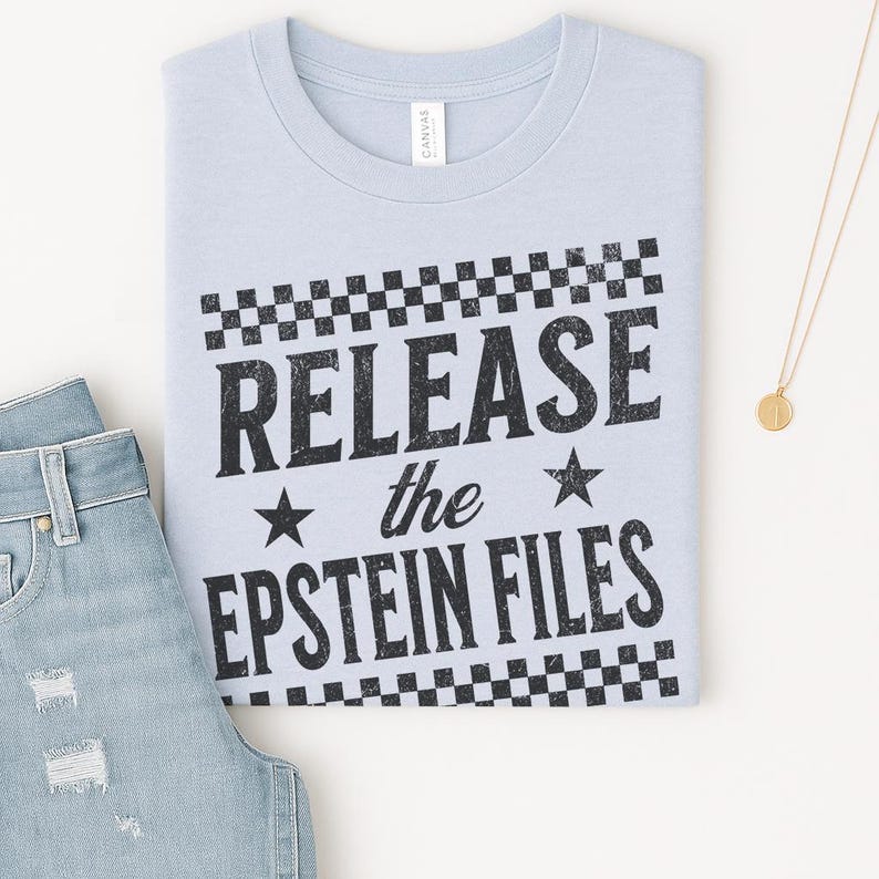 May include: A light blue t-shirt with the text "RELEASE the EPSTEIN FILES" in black, framed by a checkered pattern. The shirt is folded next to a pair of light blue jeans and a gold necklace.