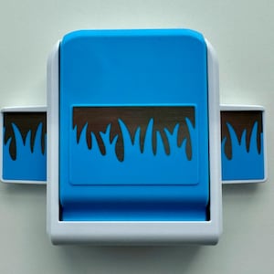 Könnte beinhalten: Ein blau-weißer Papierbastelstanzer mit einem Grasdesign. Der Stanzer hat eine rechteckige Form mit einem blauen Oberteil und einem weißen Rahmen. Das Grasdesign ist dunkelbraun.
