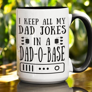 May include: White ceramic coffee mug with a black handle and black rim. The mug has a black graphic that says "I keep all my dad jokes in a dad-o-base" with a database icon and a server icon.