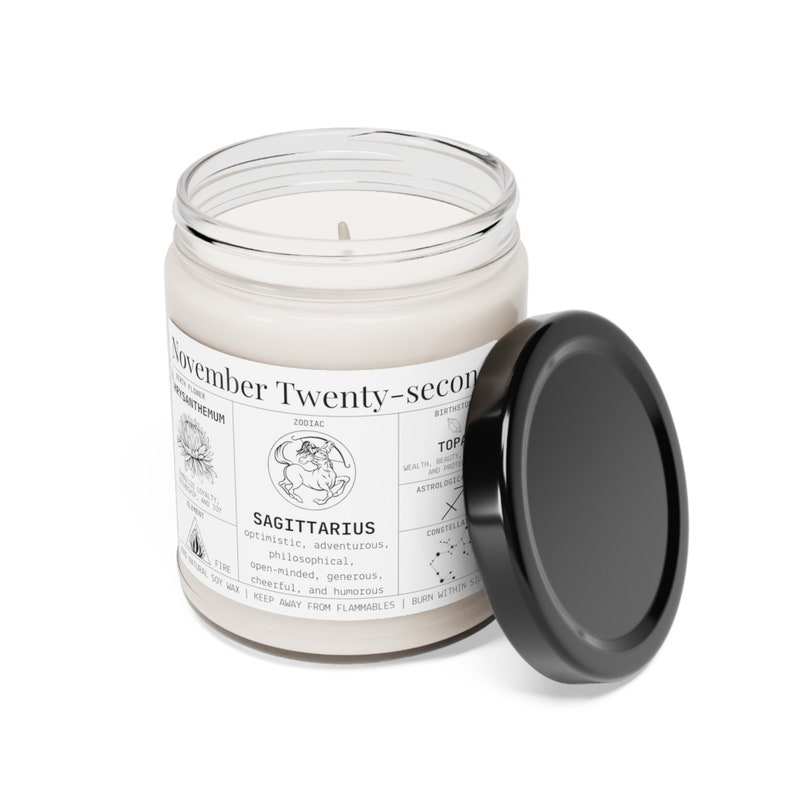 May include: A white candle in a clear glass jar with a black lid. The label reads "November Twenty-second" and features the Sagittarius zodiac sign, along with characteristics like "optimistic" and "adventurous."