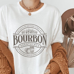 May include: White t-shirt with a brown graphic print of a circle with the text "TOO MUCH OF ANYTHING IS BAD, BUT TOO MUCH BOURBON IS BARELY ENOUGH" and "BOURBON KENTUCKY" in the center.