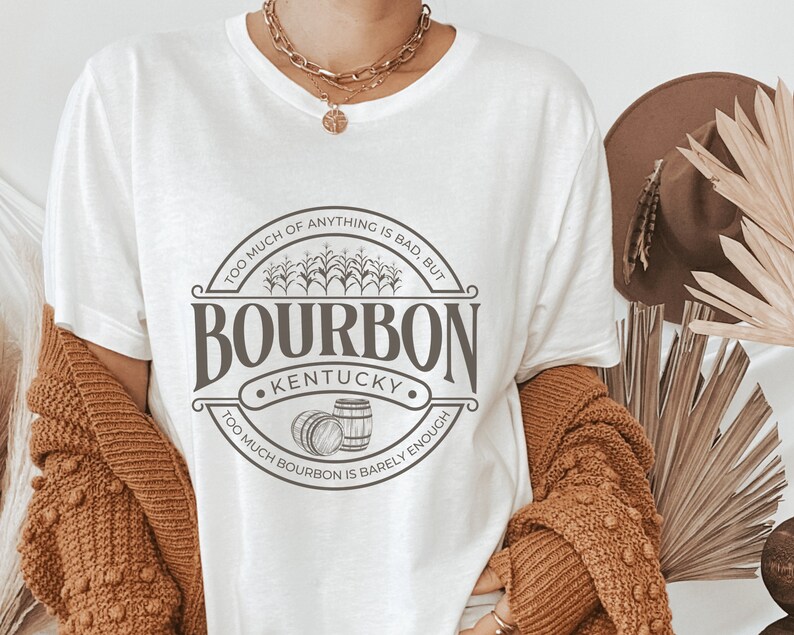 May include: White t-shirt with a brown graphic print of a circle with the text "TOO MUCH OF ANYTHING IS BAD, BUT TOO MUCH BOURBON IS BARELY ENOUGH" and "BOURBON KENTUCKY" in the center.