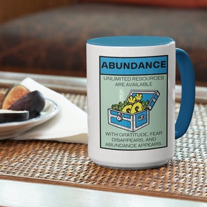 May include: White ceramic mug with a blue handle. The mug has a graphic of a treasure chest filled with money bags and the text "Abundance, Unlimited Resources Are Available. With Gratitude, Fear Disappears, and Abundance Appears."