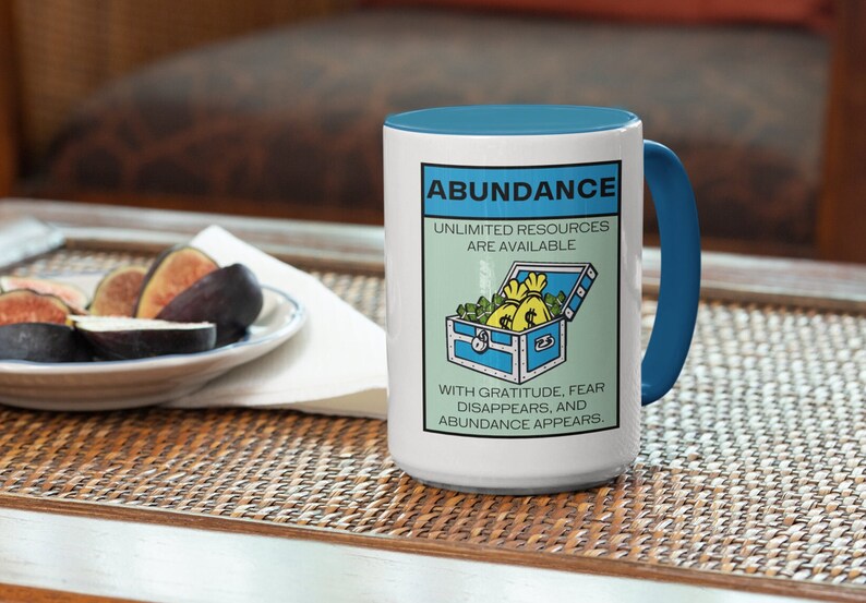 May include: White ceramic mug with a blue handle. The mug has a graphic of a treasure chest filled with money bags and the text "Abundance, Unlimited Resources Are Available. With Gratitude, Fear Disappears, and Abundance Appears."