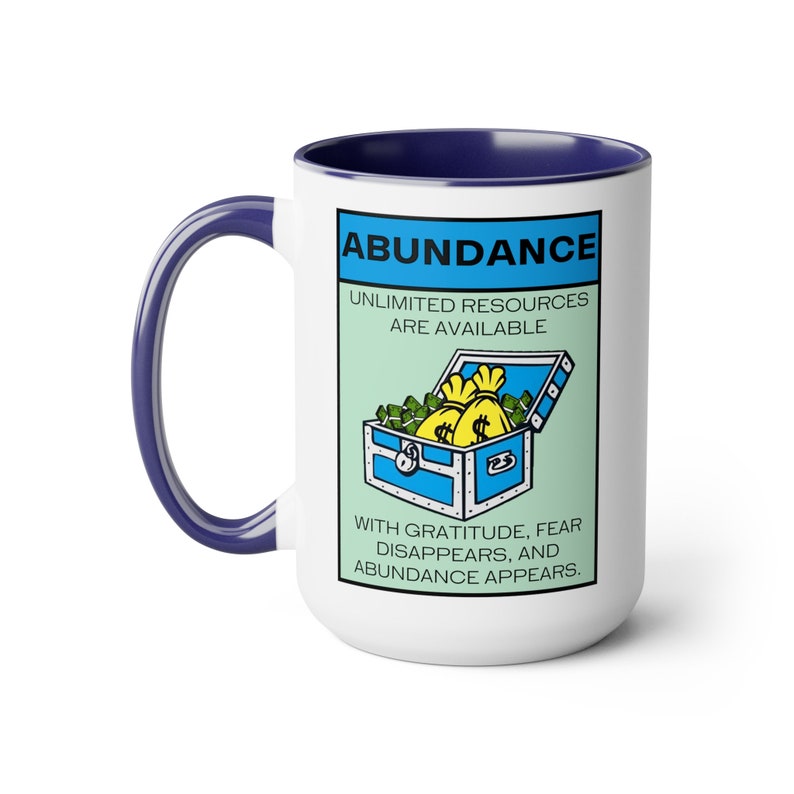 May include: White ceramic coffee mug with a blue rim. The mug features a cartoon illustration of a treasure chest overflowing with money bags and the text "ABUNDANCE, UNLIMITED RESOURCES ARE AVAILABLE, WITH GRATITUDE, FEAR DISAPPEARS, AND ABUNDANCE APPEARS."
