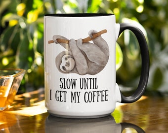 May include: White ceramic coffee mug with a black rim and handle. A cartoon sloth hangs from a branch with the text "Slow Until I Get My Coffee".