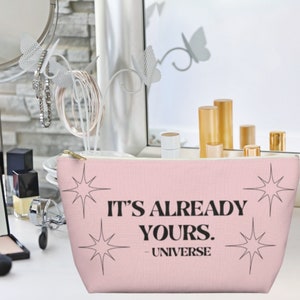 May include: Pink makeup bag with a white zipper and the text "It's already yours. - Universe" in black. The bag has a starburst design on each side.