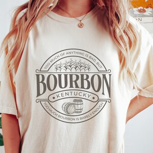 May include: A white t-shirt with a brown graphic design that says "Too much of anything is bad, but... Bourbon Kentucky... Too much bourbon is barely enough". The graphic also includes a drawing of corn stalks and two barrels.