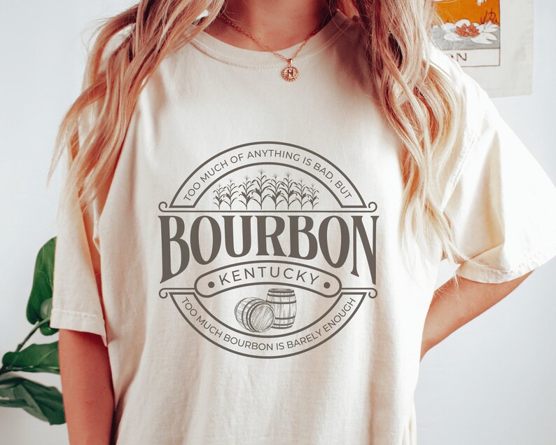 May include: A white t-shirt with a brown graphic design that says "Too much of anything is bad, but... Bourbon Kentucky... Too much bourbon is barely enough". The graphic also includes a drawing of corn stalks and two barrels.