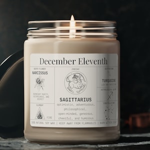 May include: A lit candle in a glass jar with a label that reads "December Eleventh" and "Sagittarius." The label includes zodiac details, birth flower, and gemstone information. The candle is made with 100% natural soy wax.