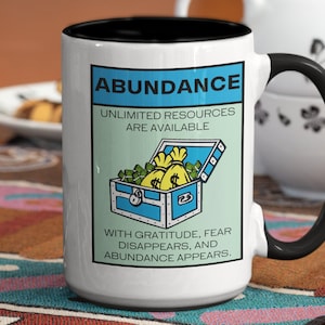 May include: White ceramic coffee mug with a black rim and handle. The mug features a graphic of a blue treasure chest overflowing with gold coins and bags. The text on the mug reads "Abundance, Unlimited Resources Are Available. With Gratitude, Fear Disappears, and Abundance Appears."