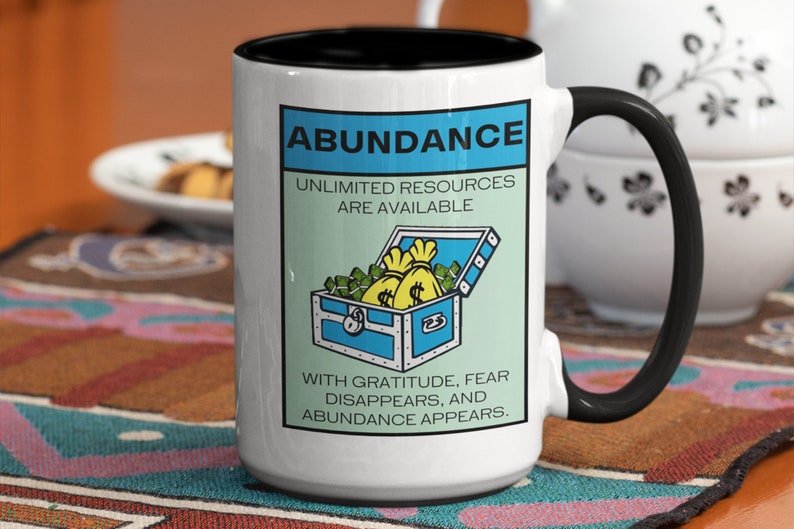 May include: White ceramic coffee mug with a black rim and handle. The mug features a graphic of a blue treasure chest overflowing with gold coins and bags. The text on the mug reads "Abundance, Unlimited Resources Are Available. With Gratitude, Fear Disappears, and Abundance Appears."