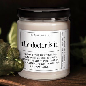 May include: A white candle with a black lid and label that reads "Ph. Done. serenity the doctor is in". The label also includes the text "Celebrate your achievement and relax after all your hard work because you didn't spend years on your dissertation just to blow out a regular candle." and "100% Natural Soy Wax | Keep Away From Flammables | Burn Within Sight".