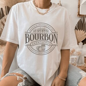 May include: White t-shirt with a brown graphic design that says "Bourbon Kentucky" with two barrels and the text "Too much of anything is bad, but too much bourbon is barely enough."