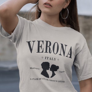 May include: A beige t-shirt with black text that reads "VERONA + ITALY + Montague A PAIR OF STAR-CROSS'D LOVERS Capulet". The text is centered on the shirt and features a silhouette of two people facing each other.