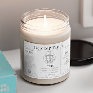 May include: A lit, beige-colored candle in a clear glass jar with a white label that reads "October Tenth" and "Libra." The label also includes the birth flower, marigold, and the zodiac sign's traits. A black lid sits nearby.