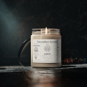 May include: A lit, beige-colored candle in a clear glass jar with a black lid. The label reads "November Second" and features zodiac information for Scorpio, including birth flower, gemstone, and traits. The candle is made with natural soy wax.