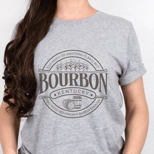 May include: Gray t-shirt with a brown graphic of a circle with the text "TOO MUCH OF ANYTHING IS BAD, BUT TOO MUCH BOURBON IS BARELY ENOUGH" and the word "BOURBON" in a large font with the text "KENTUCKY" underneath.