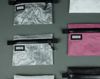 Può includere: Sei pochette rettangolari con cerniera in vari colori e texture, tra cui argento, bianco, rosa e nero. Ogni pochette presenta una cerniera orizzontale nera, una linguetta laterale nera e un'etichetta nera con il testo bianco KREIS sul davanti.