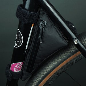 May include: A black bike frame with a black triangle-shaped handlebar bag attached to the frame. The bag has a zipper and a small pocket on the front. The bike has a brown tyre with a black tread.